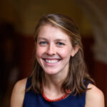 Woman with light brown shoulder length hair & blue eyes, smiling, and wearing a sleeveless blue dress and red beaded necklace